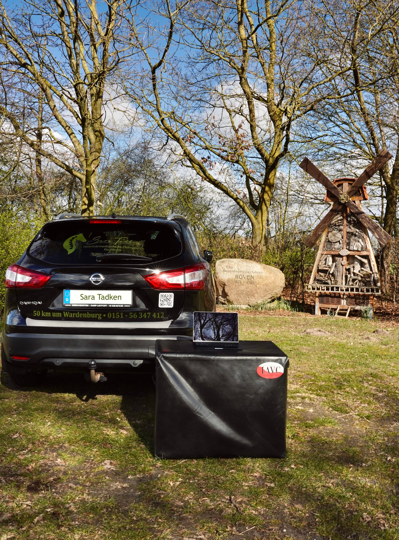 Auto mit einem schwarzen Bale vor Bäumen, einem Stein auf dem Höven steht und einer Windmühle. Sara Tadken steht auf dem Auto